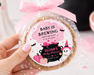 A hand holds a round baked good wrapped in clear cellophane and tied with a pink ribbon, featuring a custom round label that reads 'BABY IS BREWING' with a cartoon witch and cat. The background is a styled pink Halloween-themed table setting with cupcakes, a ghost decoration, and fairy lights.