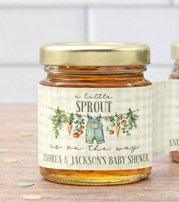 Two glass jars of honey with gold lids and personalized labels, displayed on a wooden surface with a soft-focus background.