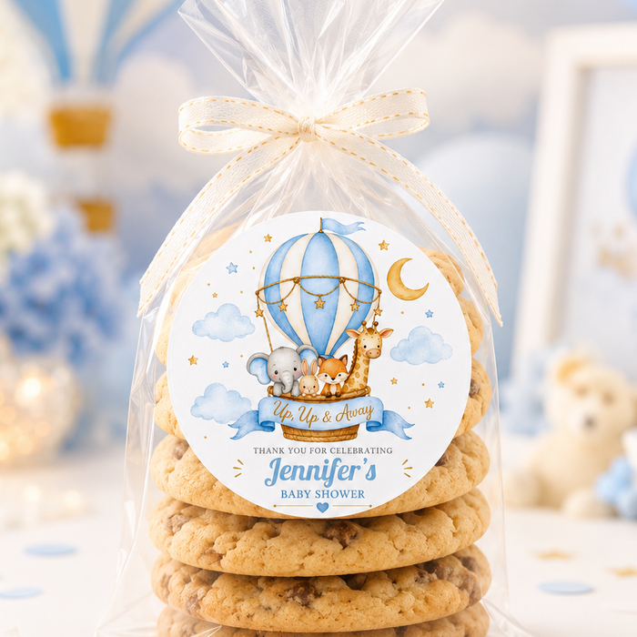 A stack of personalized chocolate chip cookies inside a clear cellophane gift bag tied with a cream ribbon, featuring a round hot air balloon baby shower label. The background is a soft, blurred blue and white themed party setting.