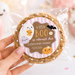 A hand holds a packaged round cookie with a Halloween-themed printed label that reads 'a little BOO is almost due' and 'Elena's Baby Shower 10.24.