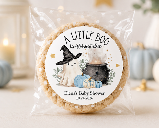 A round rice cake in a clear cellophane bag with a custom printed circular label that reads 'A LITTLE BOO is almost due' and features a witch, ghost, pumpkin, and cauldron illustration. The cake is photographed on a light surface with softly blurred decorative pumpkins, candles, and a lantern in the background.