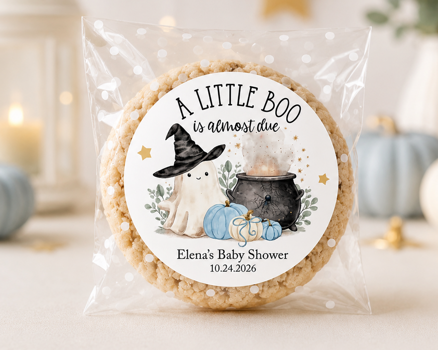 A round rice cake in a clear cellophane bag with a custom printed circular label that reads 'A LITTLE BOO is almost due' and features a witch, ghost, pumpkin, and cauldron illustration. The cake is photographed on a light surface with softly blurred decorative pumpkins, candles, and a lantern in the background.
