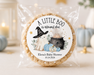 A round rice cake in a clear cellophane bag with a custom printed circular label that reads 'A LITTLE BOO is almost due' and features a witch, ghost, pumpkin, and cauldron illustration. The cake is photographed on a light surface with softly blurred decorative pumpkins, candles, and a lantern in the background.
