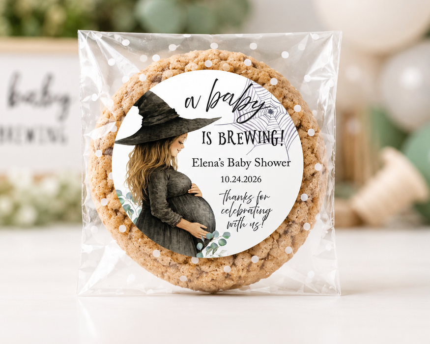 A round custom baby shower cookie favor in a clear plastic bag, featuring a printed label with a witch illustration and the text 'a baby IS BREWING! Elena's Baby Shower' on a white background with blurred decorative props.