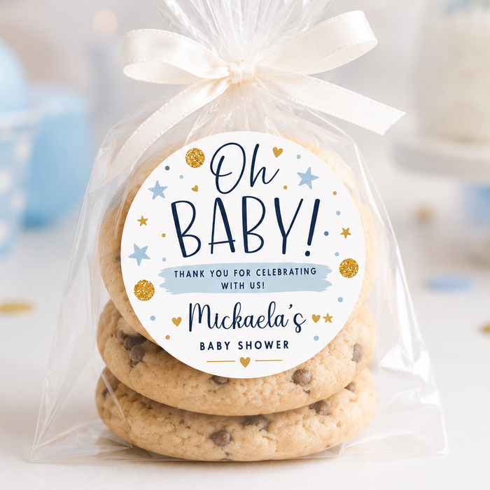 A clear cellophane bag tied with a white ribbon, containing three stacked chocolate chip cookies, with a round printed label that reads 'Oh BABY! Thank you for celebrating with us! Michaela's Baby Shower.' The scene is softly lit and decorated with pastel blue balloons and party confetti in the background.