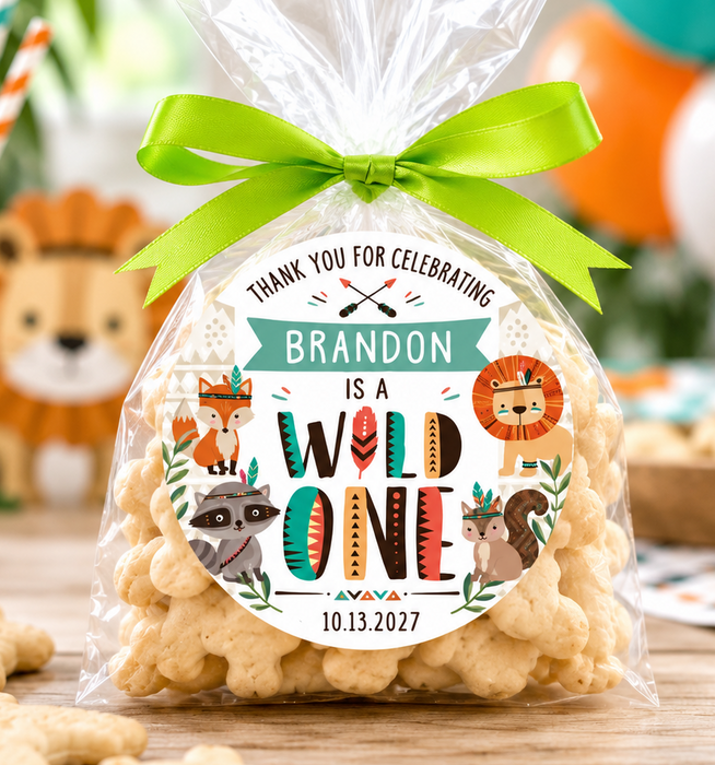 A clear cellophane bag filled with star-shaped cookies, tied with a bright green ribbon and wrapped around with a custom printed party label that reads 'BRANDON IS A WILD ONE' with animal illustrations. The bag is photographed on a wooden table with party decorations, balloons, and snacks in the background.