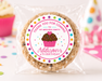A round chocolate chip cookie sealed in a clear plastic bag with a colorful round printed label that reads 'Thank you for coming to my birthday party! Addison 6th Birthday' and features a cupcake illustration. The scene is set on a white surface with blurred party decorations in the background.
