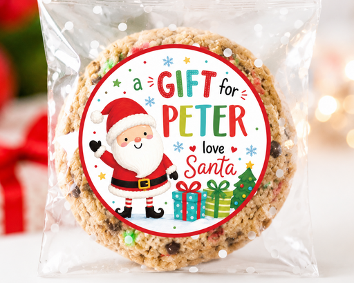 A round chocolate chip cookie sealed in a clear plastic bag with a festive circular label that reads 'a GIFT for PETER love Santa' with a cartoon Santa, Christmas tree, and gift boxes, set against a blurred holiday background.