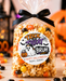 A clear cellophane bag filled with Halloween popcorn and candy, tied with a black ribbon, and labeled 'Trick or Treat Bruh' with a custom 'Happy Halloween from: Jason' note. The bag is set on a wooden surface with scattered candy corn, black candies, and blurred orange jack-o'-lanterns in the background.
