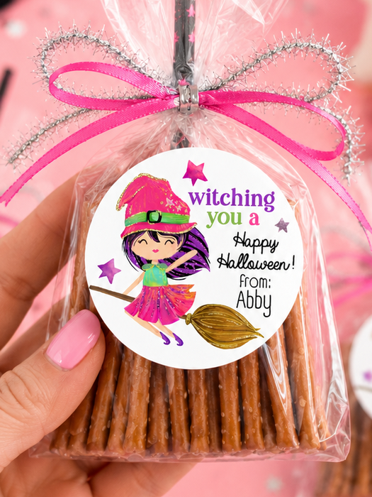 A hand holds a clear cellophane bag of pretzel sticks tied with a pink ribbon and glittery silver bow, featuring a round witch-themed label that reads 'witching you a Happy Halloween! from: Abby.' The scene is styled in pink with Halloween props including a black witch hat, ghost, pom-poms, and another packaged treat bag in the background.