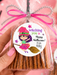 A hand holds a clear cellophane bag of pretzel sticks tied with a pink ribbon and glittery silver bow, featuring a round witch-themed label that reads 'witching you a Happy Halloween! from: Abby.' The scene is styled in pink with Halloween props including a black witch hat, ghost, pom-poms, and another packaged treat bag in the background.