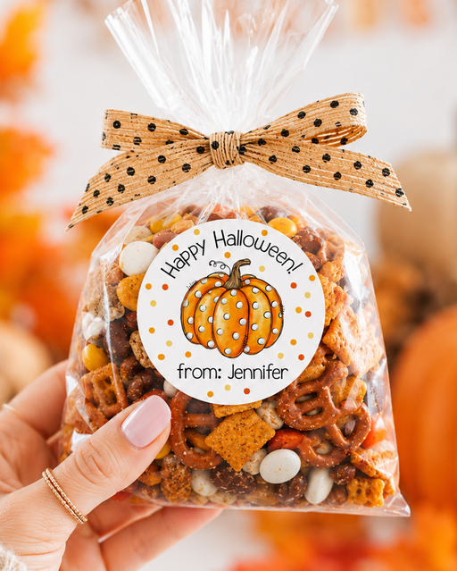 A hand holds a clear cellophane bag filled with a Halloween-themed snack mix, tied with a tan polka-dot bow. The front has a round white label with a pumpkin illustration and the text 'Happy Halloween from: Jennifer'.