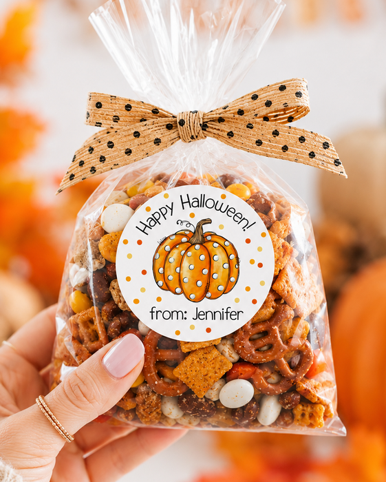 A hand holds a clear cellophane bag filled with a Halloween-themed snack mix, tied with a tan polka-dot bow. The front has a round white label with a pumpkin illustration and the text 'Happy Halloween from: Jennifer'.