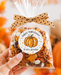 A hand holds a clear cellophane bag filled with a Halloween-themed snack mix, tied with a tan polka-dot bow. The front has a round white label with a pumpkin illustration and the text 'Happy Halloween from: Jennifer'.