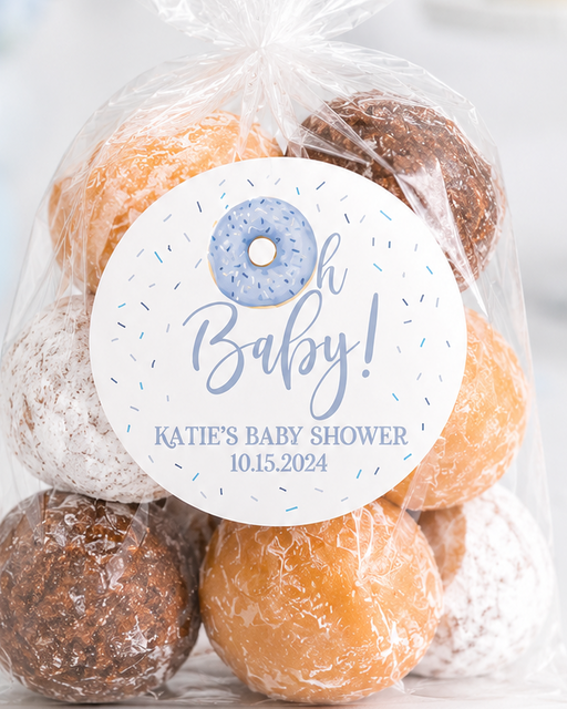 A clear cellophane bag filled with assorted donuts or doughnut-shaped treats, sealed with a twist, and topped with a round white label that reads 'Oh Baby! Katie's Baby Shower 10.15.