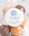 A clear cellophane bag filled with assorted donuts or doughnut-shaped treats, sealed with a twist, and topped with a round white label that reads 'Oh Baby! Katie's Baby Shower 10.15.