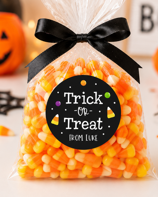 A clear cellophane bag filled with orange, yellow, and white candy corn, tied with a black ribbon and featuring a round black label that reads 'Trick or Treat From Luke.' The bag is photographed on a white surface with Halloween decorations in the background, including a jack-o'-lantern, black spiderwebs, orange pom-poms, and a sign reading BOO.