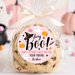 A clear cellophane bag tied with a cream ribbon, containing chocolate chip cookies, with a round pink 'hey Boo! Halloween treat just for you' label on the front. The scene is styled with Halloween props including pumpkins, a ghost, and candy in the background.