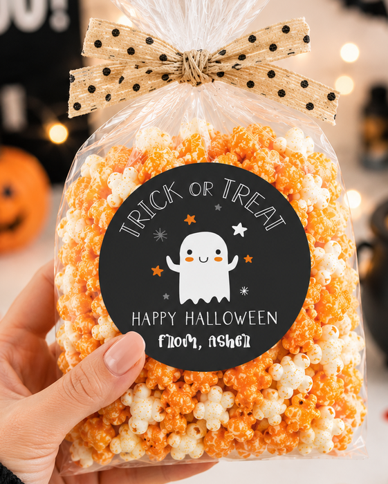 A hand holds a clear cellophane bag of Halloween popcorn tied with a beige polka-dot bow. The bag has a round black label that says 'Trick or Treat Happy Halloween Noah, 9yo'.