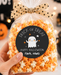 A hand holds a clear cellophane bag of Halloween popcorn tied with a beige polka-dot bow. The bag has a round black label that says 'Trick or Treat Happy Halloween Noah, 9yo'.