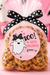 A clear cellophane bag filled with chocolate chip cookies, tied with a black polka-dot bow, and topped with a round pink label that says 'BOO! Happy Halloween from: Victoria.' The scene is styled in pink with Halloween decorations in the background.
