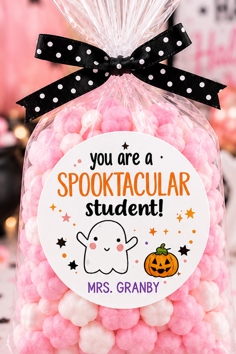 A clear cellophane bag filled with pink and white pom-pom candies, tied with a black polka-dot ribbon, and featuring a round label that reads 'you are a Spooktacular student! Mrs. Granby.