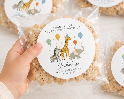 A hand holds a clear bag of personalized rice crispy cookies with a round animal-themed party label, surrounded by five similar bags on a light wood table with party props.