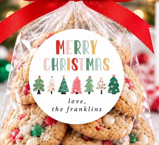 A clear cellophane gift bag filled with Christmas cookies and tied with a bright red ribbon, featuring a round white label that reads 'MERRY CHRISTMAS love, the franklins' with colorful tree illustrations. The bag is photographed on a marble surface with a blurred festive holiday background including lights, ornaments, a gingerbread house, and a mug that says 'Merry Christmas'.