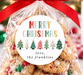 A clear cellophane gift bag filled with Christmas cookies and tied with a bright red ribbon, featuring a round white label that reads 'MERRY CHRISTMAS love, the franklins' with colorful tree illustrations. The bag is photographed on a marble surface with a blurred festive holiday background including lights, ornaments, a gingerbread house, and a mug that says 'Merry Christmas'.