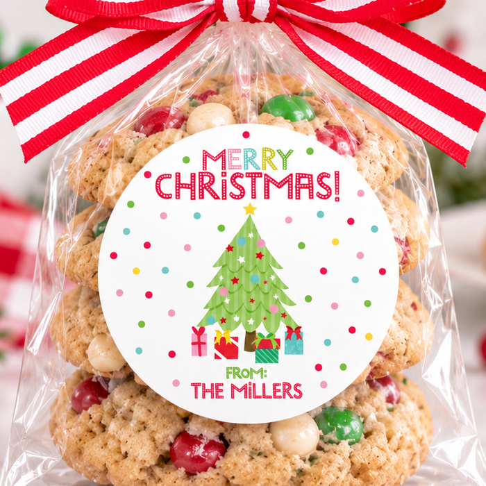 A clear cellophane gift bag filled with Christmas cookies and tied with a red-and-white striped ribbon, featuring a round 'Merry Christmas From The Millers' label. The bag is photographed on a marble surface with festive holiday decorations in the background.