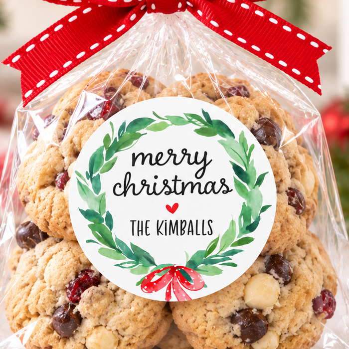 A bag of holiday cookies tied with a red ribbon and a round 'merry christmas' label, set on a marble surface with a blurred Christmas tree and ornaments in the background.