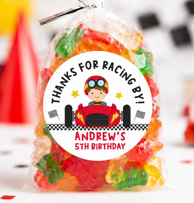 A clear cellophane bag filled with colorful gummy candies and tied with a clear twist tie, featuring a round racing-themed label that reads 'THANKS FOR RACING BY! ANDREW'S 5TH BIRTHDAY' with a cartoon driver in a red race car. The bag is photographed on a white party surface with blurred balloons, checkered backdrop elements, and toy cars in the background.
