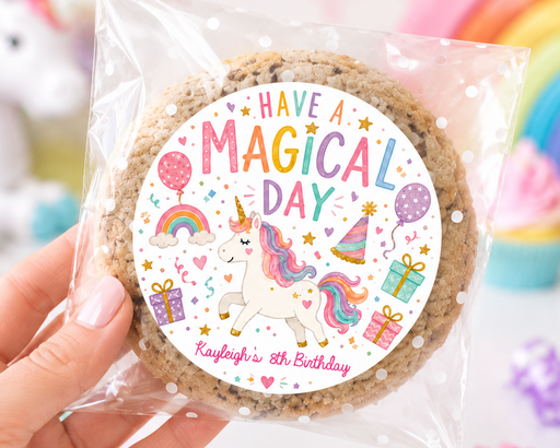 A hand holds a clear plastic bag containing a round chocolate chip cookie with a colorful custom sticker that reads 'HAVE A MAGICAL DAY' and 'Kayleigh's 6th Birthday' with a unicorn illustration. The background is a soft-focus pastel scene with a rainbow, cupcakes, and a unicorn.