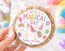 A hand holds a clear plastic bag containing a round chocolate chip cookie with a colorful custom sticker that reads 'HAVE A MAGICAL DAY' and 'Kayleigh's 6th Birthday' with a unicorn illustration. The background is a soft-focus pastel scene with a rainbow, cupcakes, and a unicorn.
