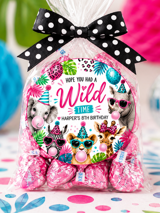 A clear cellophane bag of wrapped pink chocolates tied with a black-and-white polka dot bow, featuring a colorful round label with cartoon animals and the text 'Hope You Had A Wild 8th Birthday.' The bag is photographed on a decorated surface with tropical leaves and bright party props in the background.