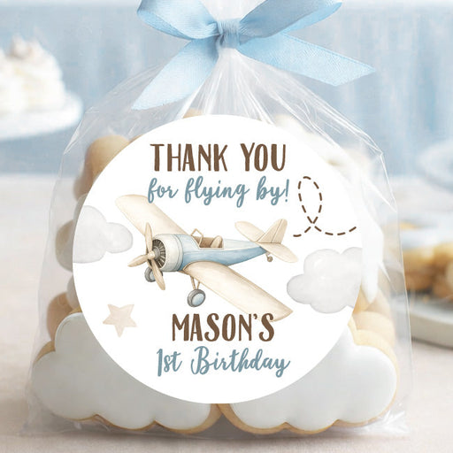 A clear cellophane favor bag tied with a light blue ribbon, filled with cloud-shaped cookies and featuring a round printed label that reads 'THANK YOU for flying by! MASON'S 1st Birthday' with an airplane illustration. The bag is photographed on a tabletop with a softly blurred party background in pastel blue tones.