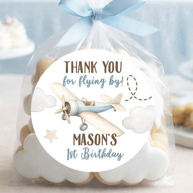 A clear cellophane favor bag tied with a light blue ribbon, filled with cloud-shaped cookies and featuring a round printed label that reads 'THANK YOU for flying by! MASON'S 1st Birthday' with an airplane illustration. The bag is photographed on a tabletop with a softly blurred party background in pastel blue tones.