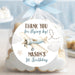 A clear cellophane favor bag tied with a light blue ribbon, filled with cloud-shaped cookies and featuring a round printed label that reads 'THANK YOU for flying by! MASON'S 1st Birthday' with an airplane illustration. The bag is photographed on a tabletop with a softly blurred party background in pastel blue tones.