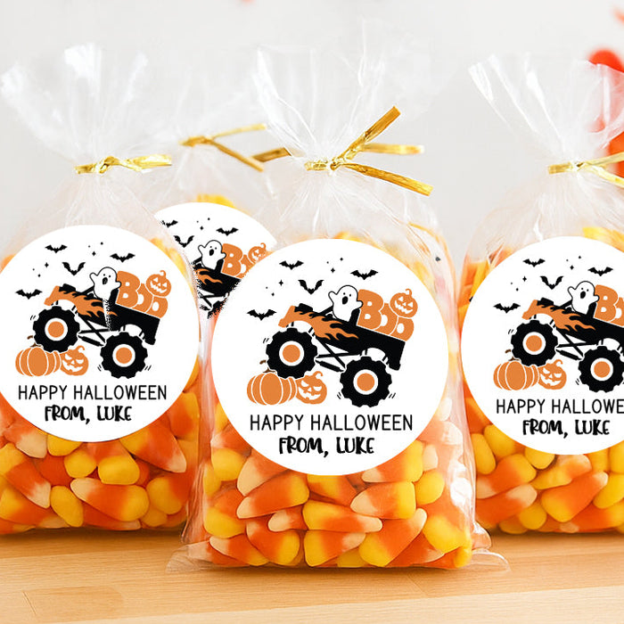 Three clear plastic bags filled with candy corn are tied with gold ribbons and arranged on a wooden surface. Each bag has a round white label featuring an orange monster truck, pumpkins, bats, and the text 'HAPPY HALLOWEEN FROM, LIKE'.