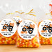 Three clear plastic bags filled with candy corn are tied with gold ribbons and arranged on a wooden surface. Each bag has a round white label featuring an orange monster truck, pumpkins, bats, and the text 'HAPPY HALLOWEEN FROM, LIKE'.