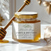 A glass jar of honey with a gold lid and a white personalized label reading 'Chris & Brittany' and 'June 15, 2027', styled on a white surface with a honey dipper and soft beige fabric in a bright, elegant setting.